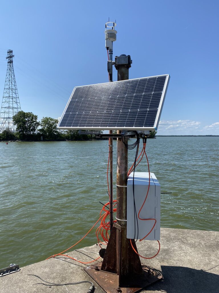 Near the water's edge a solar panel and a grey box are attached to a pole. 