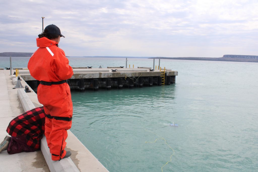 A person in an orange jumpsuit looks off a dock, testdriving an ROV tethered with a yellow cord.