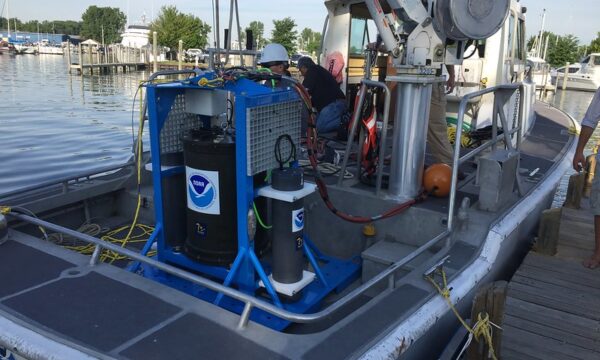 A large metal barrel-like device sits on the deck of a boat with a NOAA logo.