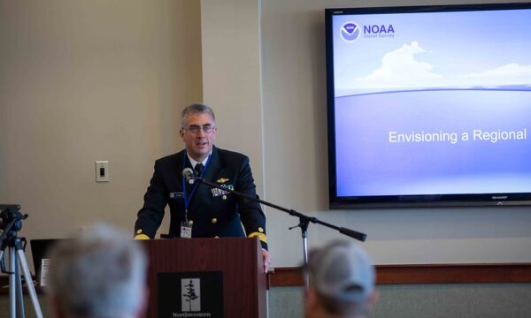 A person in a uniform speaks at a podium in front of a NOAA slide