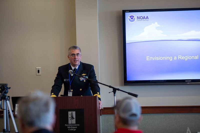 A person in a uniform speaks at a podium in front of a NOAA slide